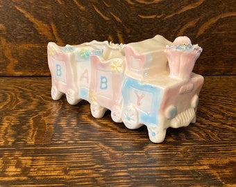 May include: A ceramic baby train painted in pink, blue and white. Each carriage is shaped like a block with a letter, spelling out "BABY".