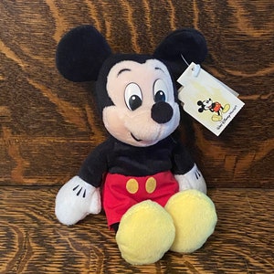 May include: A plush Mickey Mouse toy wearing red shorts with yellow buttons and a black shirt. The toy has a white tag attached to its right ear that says "Walt Disney World&reg;".