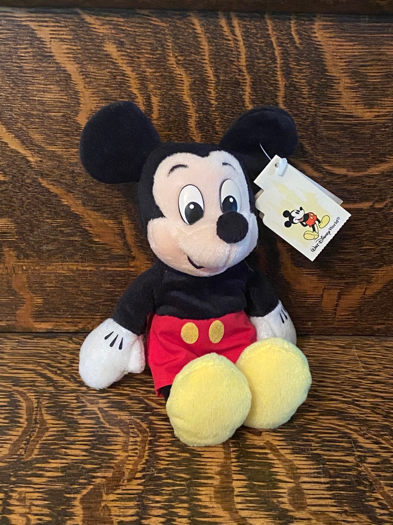 May include: A plush Mickey Mouse toy wearing red shorts with yellow buttons and a black shirt. The toy has a white tag attached to its right ear that says "Walt Disney World&reg;".