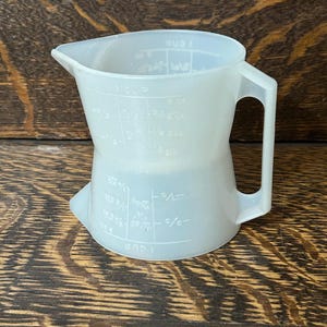 May include: A translucent white plastic measuring jug with a handle and a pouring spout. The jug has measurement markings in cups, fluid ounces, and fractions. The jug is on a wooden surface.
