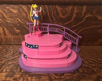 May include: A pink and purple toy stage with a miniature figurine of a character with blonde hair, wearing a sailor uniform. The stage has multiple levels, a bridge, and star and moon accents. The figurine is standing on the top level.