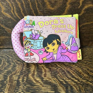 Peut inclure: Un livre en tissu intitulé Dora's Dress Up Adventure représentant Dora l'exploratrice en robe violette. La couverture comprend une poignée rose à pois, un coffre au trésor bleu, un petit personnage chevalier et un bouton sonore.