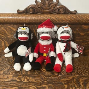 May include: Three plush sock monkey toys featuring red mouths and white heels. The left monkey wears a black and white penguin hood. The center monkey wears a red Santa suit with a white trim hat. The right monkey wears a white snowman hood and scarf.