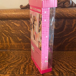 May include: A pink and white Barbie Fashion Fever doll box with a clear plastic window showing a doll with long blonde hair wearing a white dress. The box has a pink and white polka dot pattern and the text "Barbie Fashion Fever" is printed on the front.