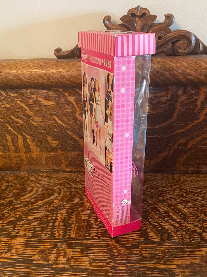 May include: A pink and white Barbie Fashion Fever doll box with a clear plastic window showing a doll with long blonde hair wearing a white dress. The box has a pink and white polka dot pattern and the text "Barbie Fashion Fever" is printed on the front.