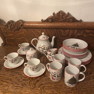 May include: A white Christmas-themed tea set with red trim. The set includes a teapot, cups, saucers, plates, a sugar bowl, and a creamer. Each piece features a winter village scene and a Christmas tree.