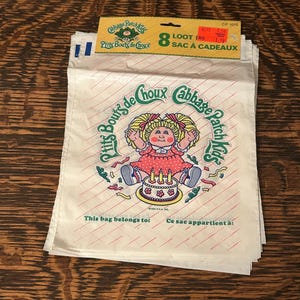 May include: White Cabbage Patch Kids loot bags with a red and white striped pattern. The bags feature a Cabbage Patch Kids graphic and text in English and French. The bags are stacked on a wooden surface.
