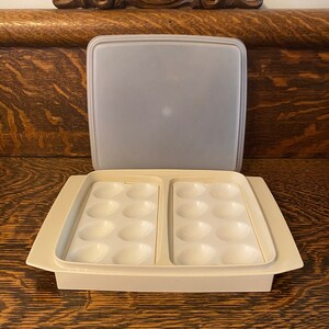 Könnte beinhalten: Ein weißer Eierkocher aus Kunststoff mit einem durchsichtigen Deckel. Der Kocher hat zwei Abschnitte, jeder mit sechs Vertiefungen für Eier. Der Deckel trägt die Aufschrift "Tupperware".