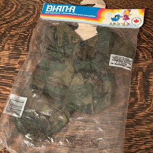 Può includere: Una confezione sigillata contenente abiti per bambole con un motivo mimetico. La confezione presenta il marchio "DIANA" e il testo "doll fashions - nouveautés pour poupées". La confezione include anche il testo "CABBAGE PATCH 16" RAGGEDY ANN".