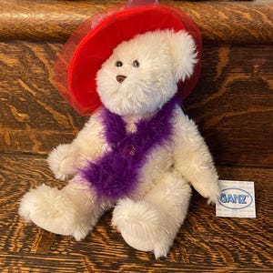 Könnte beinhalten: Weißer Plüschteddybär, der einen roten Hut mit einem Tüllschleier und einem lila Federboa trägt. Der Bär hat schwarze Augen und eine rosa Nase. Der Bär sitzt auf einer Holzoberfläche. Der Bär hat ein Etikett mit der Aufschrift "Ganz".