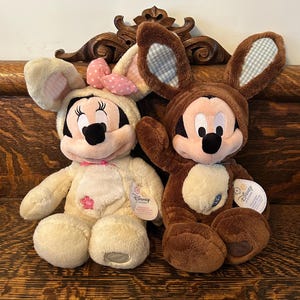 May include: Two plush Easter bunny toys, one brown and one cream, both dressed as Mickey Mouse and Minnie Mouse. The toys are sitting on a wooden surface.