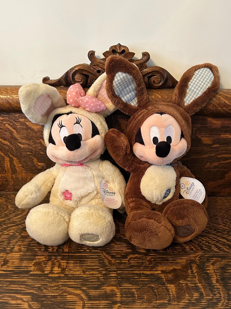 May include: Two plush Easter bunny toys, one brown and one cream, both dressed as Mickey Mouse and Minnie Mouse. The toys are sitting on a wooden surface.