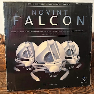 May include: A black box with the text "NOVINT FALCON" and three white and gray devices. The box also has the text "EXPERIENCE NEXT GENERATION PC GAMING" and "-IGN, BEST OF E3 GEAR."