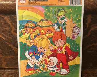 Puede incluir: Un rompecabezas vintage Rainbow Brite con marco. El rompecabezas presenta ilustraciones coloridas de personajes de Rainbow Brite, un arcoíris y casas caprichosas. El rompecabezas está diseñado para niños de 3 a 7 años. La caja tiene el logotipo de Golden y el texto "Frame-Tray. Puzzle".