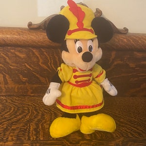 May include: A plush toy of Minnie Mouse dressed as a bandleader. She is wearing a yellow dress with red trim and a red and yellow hat with a red feather.