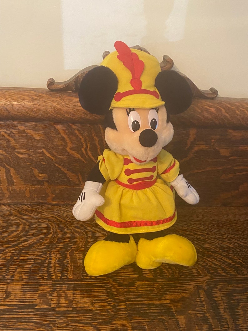 May include: A plush toy of Minnie Mouse dressed as a bandleader. She is wearing a yellow dress with red trim and a red and yellow hat with a red feather.