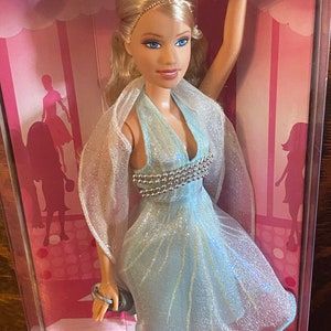 May include: A Barbie doll wearing a light blue dress with silver embellishments and a sheer white shawl. The doll has blonde hair and blue eyes. The doll is packaged in a pink box with the word "Barbie" printed on it.