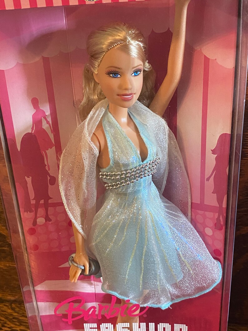 May include: A Barbie doll wearing a light blue dress with silver embellishments and a sheer white shawl. The doll has blonde hair and blue eyes. The doll is packaged in a pink box with the word "Barbie" printed on it.