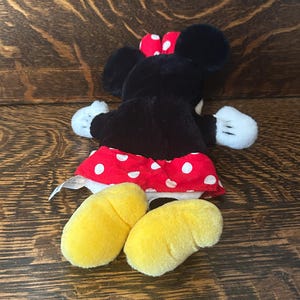 Pu&ograve; includere: Un peluche di un topo dei cartoni animati, con pelliccia nera, guanti bianchi e piedi gialli. Il topo indossa una gonna rossa a pois bianchi e un fiocco rosso a pois bianchi. Il giocattolo &egrave; appoggiato su una superficie di legno.