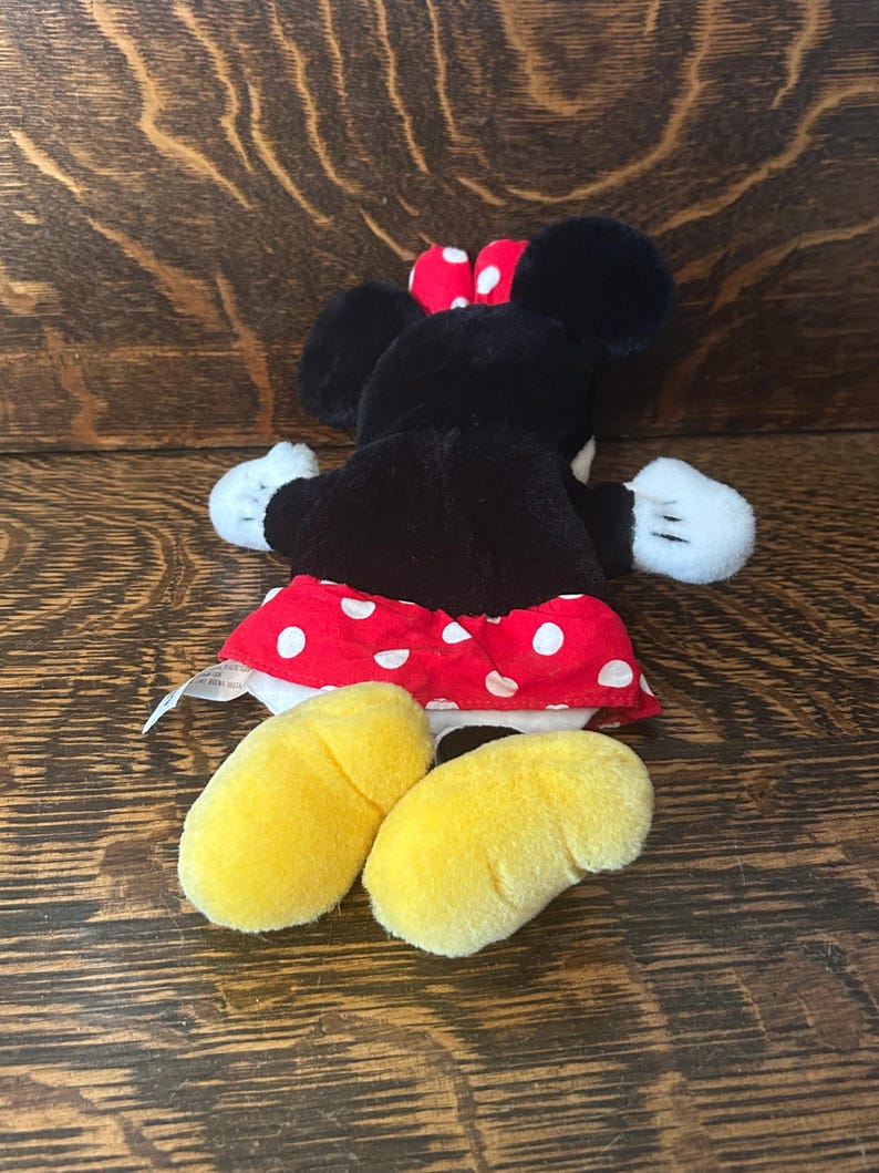 Pu&ograve; includere: Un peluche di un topo dei cartoni animati, con pelliccia nera, guanti bianchi e piedi gialli. Il topo indossa una gonna rossa a pois bianchi e un fiocco rosso a pois bianchi. Il giocattolo &egrave; appoggiato su una superficie di legno.