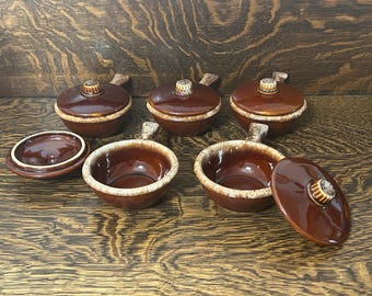 Puede incluir: Un juego de seis cuencos de cerámica marrón con asas y tapas a juego. Cada cuenco presenta un esmalte marrón oscuro brillante con un borde texturizado de color marrón más claro. Las tapas tienen un pequeño pomo estriado en el centro.
