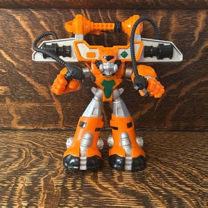 May include: An orange and silver robot toy with black accents. The toy has a lion-like face, a green chest piece, and a large weapon on its back. The robot's legs and arms are articulated, and it stands on a wooden surface.