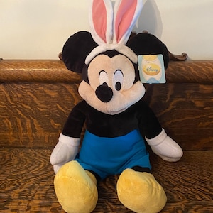 May include: A plush Mickey Mouse toy wearing pink bunny ears. The toy is sitting on a wooden surface and has a blue and black outfit. The toy has a Disney tag attached to it.
