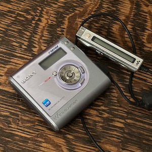 Könnte beinhalten: Ein silberner Sony MD Walkman MZ-NHF800 mit passender Fernbedienung. Das Gerät verfügt über einen UKW/MW-Tuner und ein zentrales Steuerwahlrad. Die Fernbedienung ist über ein schwarzes Kabel verbunden. Das Gerät befindet sich auf einer Holzoberfläche.