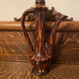 May include: A brown and white ceramic teapot with a tall, slender body and a lid with a knob. The teapot has a spout and two handles.