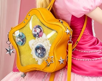 Può includere: Uno zaino giallo a forma di stella con un pannello frontale trasparente che mostra spille colorate e un dettaglio in pizzo bianco. La borsa ha una lunga tracolla regolabile e ciondoli argentati a forma di stella. Lo sfondo è rosa.