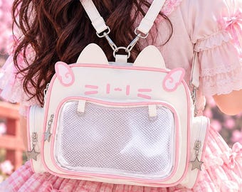 May include: A white and light pink cat-shaped backpack featuring a clear front display window, two small cat ears on top, and star-shaped zipper pulls. The bag has adjustable white straps and side pockets, designed with a clean, structured aesthetic.