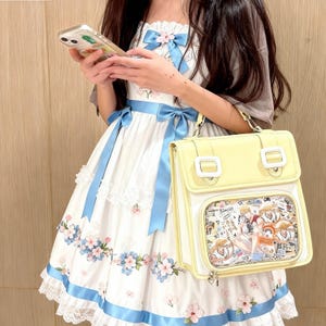 May include: A white dress with blue ribbons and floral patterns, paired with a pale yellow satchel bag. The bag has a clear front panel displaying a colorful illustration. The dress features lace trim and a bow at the neckline.