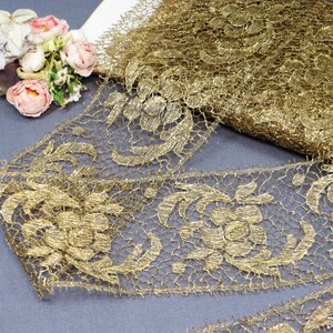 May include: Close-up of a roll of gold lace with a floral pattern. The lace is intricately designed with a delicate, open weave. A small bouquet of pink and white flowers rests on the left side of the image, adding a touch of color.