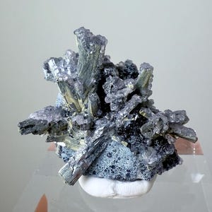 May include: A mineral specimen with a cluster of dark grey, blue-grey, and light purple crystals. The crystals have a spiky, textured appearance, with some sections appearing metallic. The mineral sits on a white base.