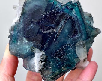May include: A cluster of blue-green fluorite crystals with a hint of purple. The crystals are intergrown and have a glassy lustre. Some of the crystals have a slight white coating.