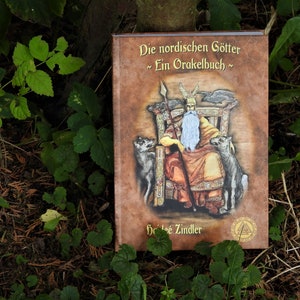 Può includere: Un libro con copertina rigida intitolato "Die nordischen Götter ~ Ein Orakelbuch ~" con un'illustrazione di una figura seduta con una lunga barba bianca, che tiene una lancia, affiancata da due lupi. Il libro ha una copertina marrone.