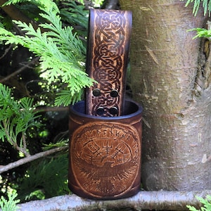 Könnte beinhalten: Ein brauner Leder-Becherhalter mit einem keltischen Knotendesign und einem Vogel mit Runen drumherum. Der Halter ist an einem braunen Lederriemen mit einem keltischen Knotendesign befestigt.