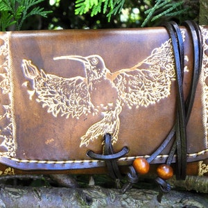 Könnte beinhalten: Eine braune Lederbörse mit einem Kolibri-Design und floralen Akzenten. Die Börse hat einen Klappverschluss und einen braunen Lederriemen mit Holzperlen.