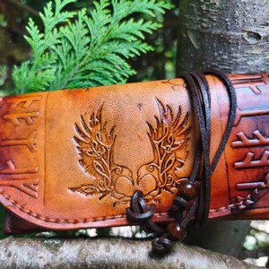 Op de afbeelding: Een beige leren etui met een in reliëf aangebracht symmetrisch vleugelontwerp in het midden en runentekens langs de zijkanten. Het etui wordt gesloten met een donkerbruin leren koord met twee kleine houten kralen aan het uiteinde.