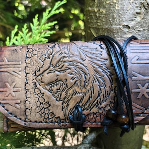Op de afbeelding: Een bruin lederen etui met een reliëf van een wolvenkop op de voorflap, omringd door uitgesneden runensymbolen. Het etui wordt gesloten met zwarte leren koorden die om de buitenkant zijn gewikkeld en zijn afgewerkt met donkere houten kralen.