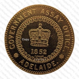May include: A gold-colored coin with the text "GOVERNMENT ASSAY OFFICE" and "ADELAIDE" around the edge. The center features a crown above the date "1852".