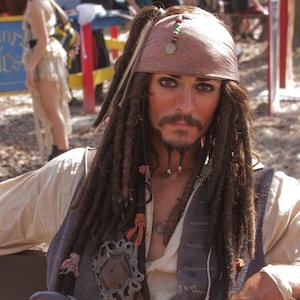 Pode incluir: Uma pessoa vestida de pirata, usando uma bandana marrom, dreadlocks e um colete sobre uma camisa branca. O traje inclui um cinto com fivela decorativa, uma pistola e anéis. O tema geral é uma fantasia de pirata.