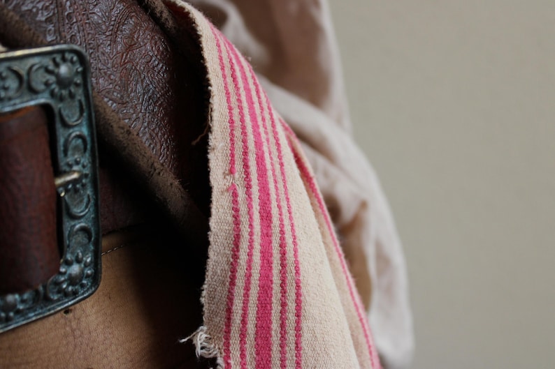 May include: A close-up of a brown leather belt with a metal buckle. The belt is worn and has a faded look. A white and pink striped fabric is draped over the belt.