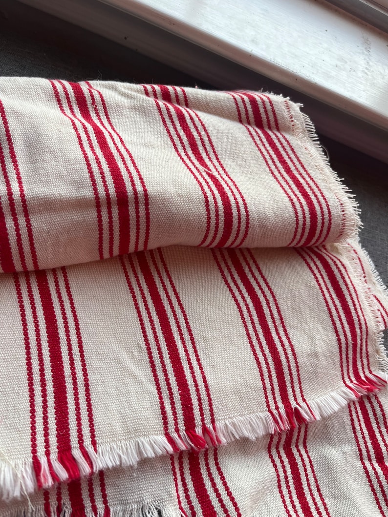 May include: White fabric with red stripes and a frayed edge. The fabric is folded in half.