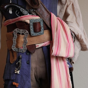 May include: A close-up of a pirate costume with a brown leather belt, silver buckles, and a striped scarf. The costume includes a blue shirt, brown pants, and a pistol.