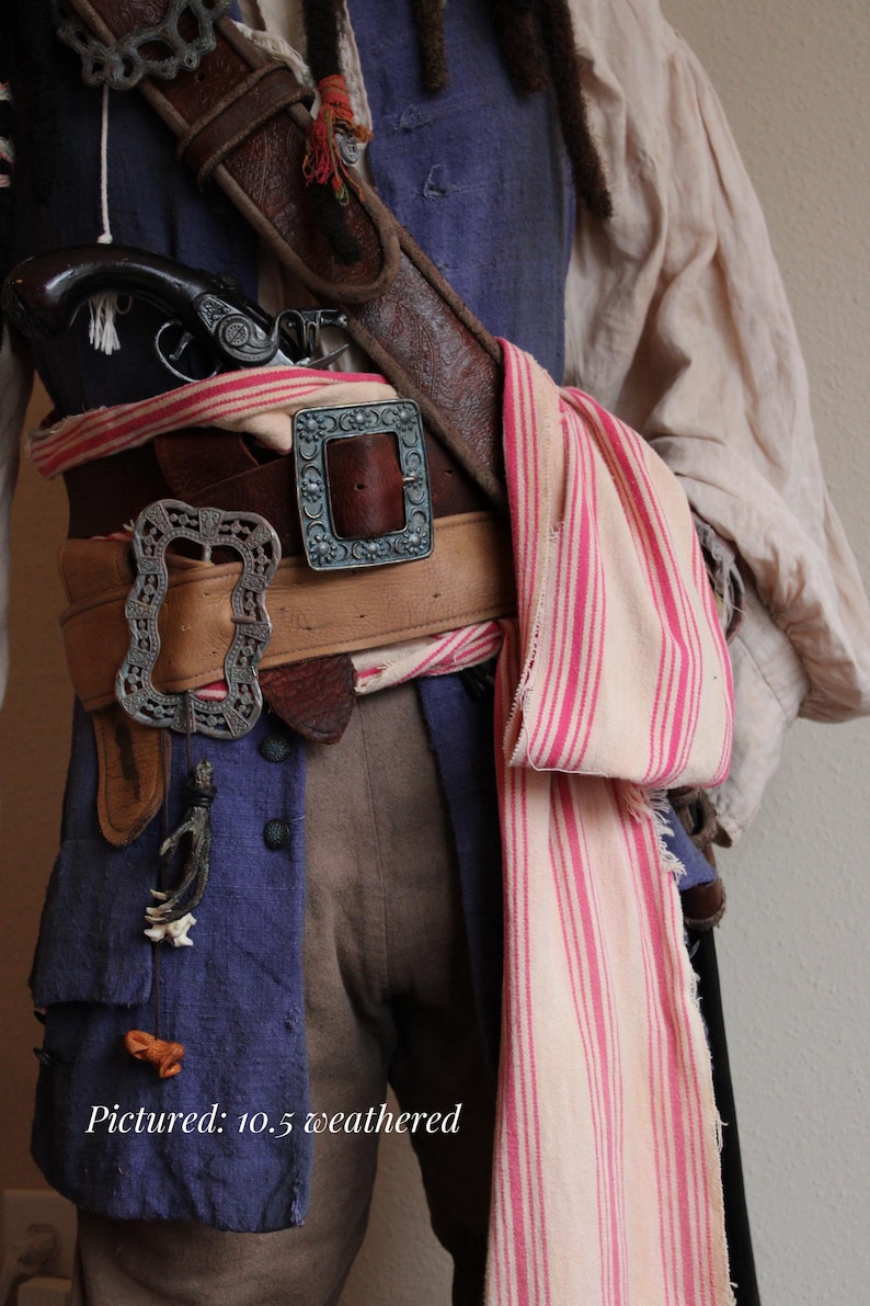 May include: A close-up of a pirate costume with a brown leather belt, silver buckles, and a striped scarf. The costume includes a blue shirt, brown pants, and a pistol.