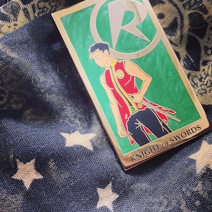 May include: A gold-colored enamel pin featuring a character with a red and black costume holding a sword. The character is standing in front of a green background with a large silver letter "R" in a circle. The pin reads "Knight of Swords".