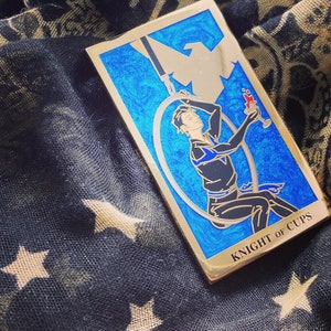 May include: A gold-colored enamel pin depicting the Knight of Cups tarot card. The card features a blue background with a swirling pattern and a figure holding a cup. The text "KNIGHT OF CUPS" is printed in gold at the bottom of the card.
