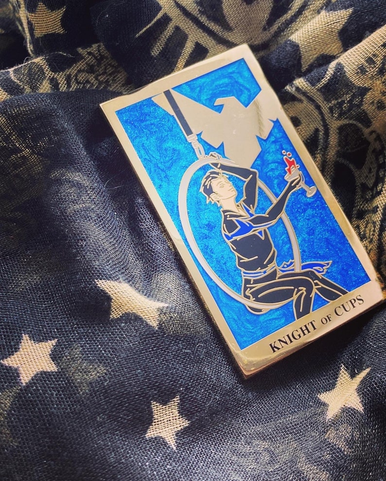 May include: A gold-colored enamel pin depicting the Knight of Cups tarot card. The card features a blue background with a swirling pattern and a figure holding a cup. The text "KNIGHT OF CUPS" is printed in gold at the bottom of the card.