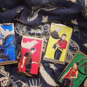 May include: Four enamel pins featuring illustrations of tarot cards. The cards are Knight of Pentacles, Knight of Wands, Knight of Swords, and Knight of Cups. The pins are gold-colored with colorful illustrations.
